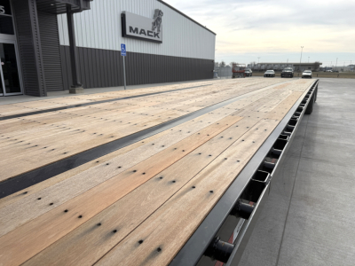 Drop deck trailer with wooden deck and metal edge rails at Mack dealership showing long platform and wheel wells.
