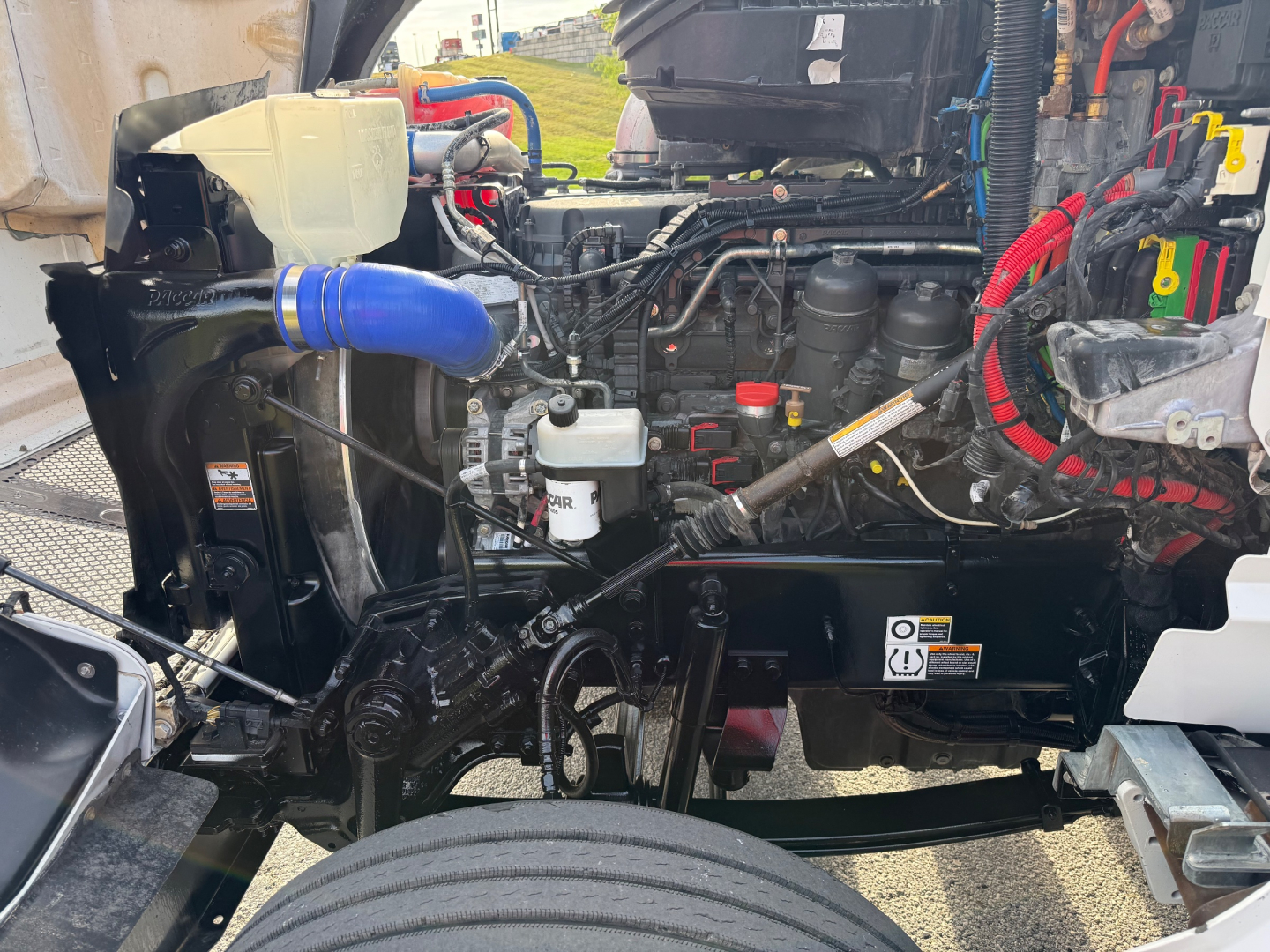 Kenworth T880S engine bay with visible coolant reservoir, blue silicone hose, air intake lines, and electrical harnesses in a truck chassis.