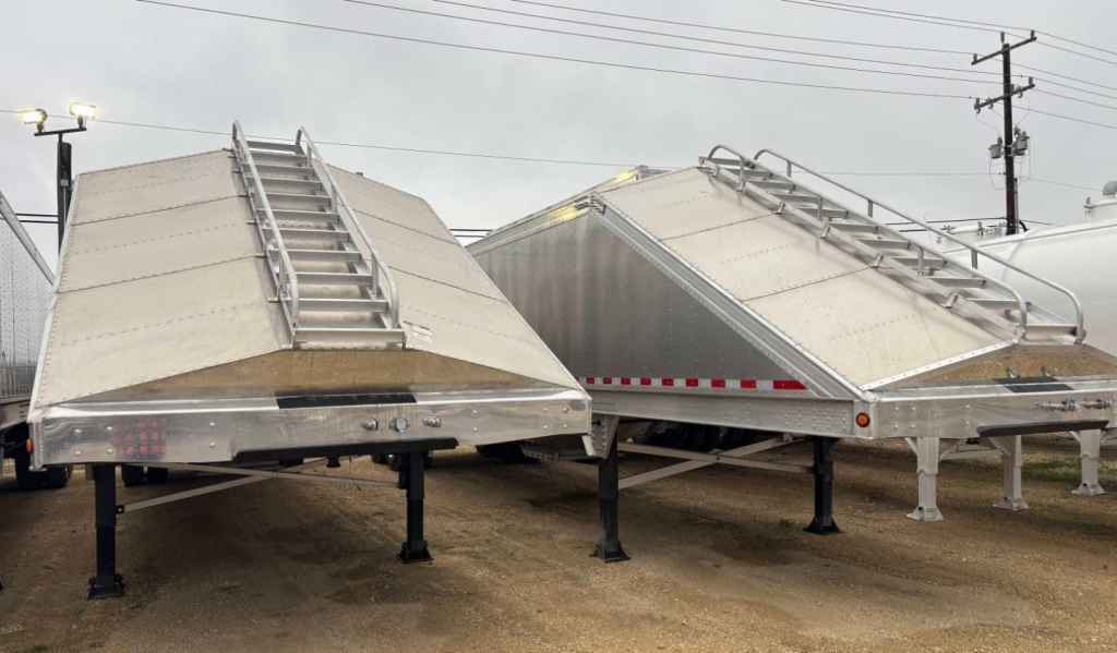Two aluminum sand/grain hopper trailers on support legs with ladders on top and ribbed sides, showing no visible branding text.