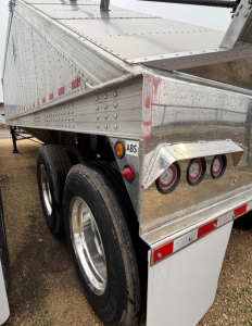 Trailer wheel area and rear corner of a new Aldura aluminum hopper trailer showing triple rear lights, reflective markers, and riveted aluminum panels.