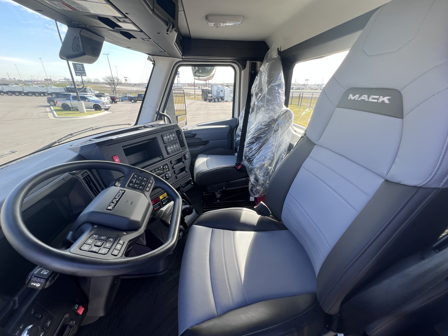 Truck cab interior of a 2027 Mack AN64T showing steering wheel, driver seat, and dashboard with center console and gear shifter.