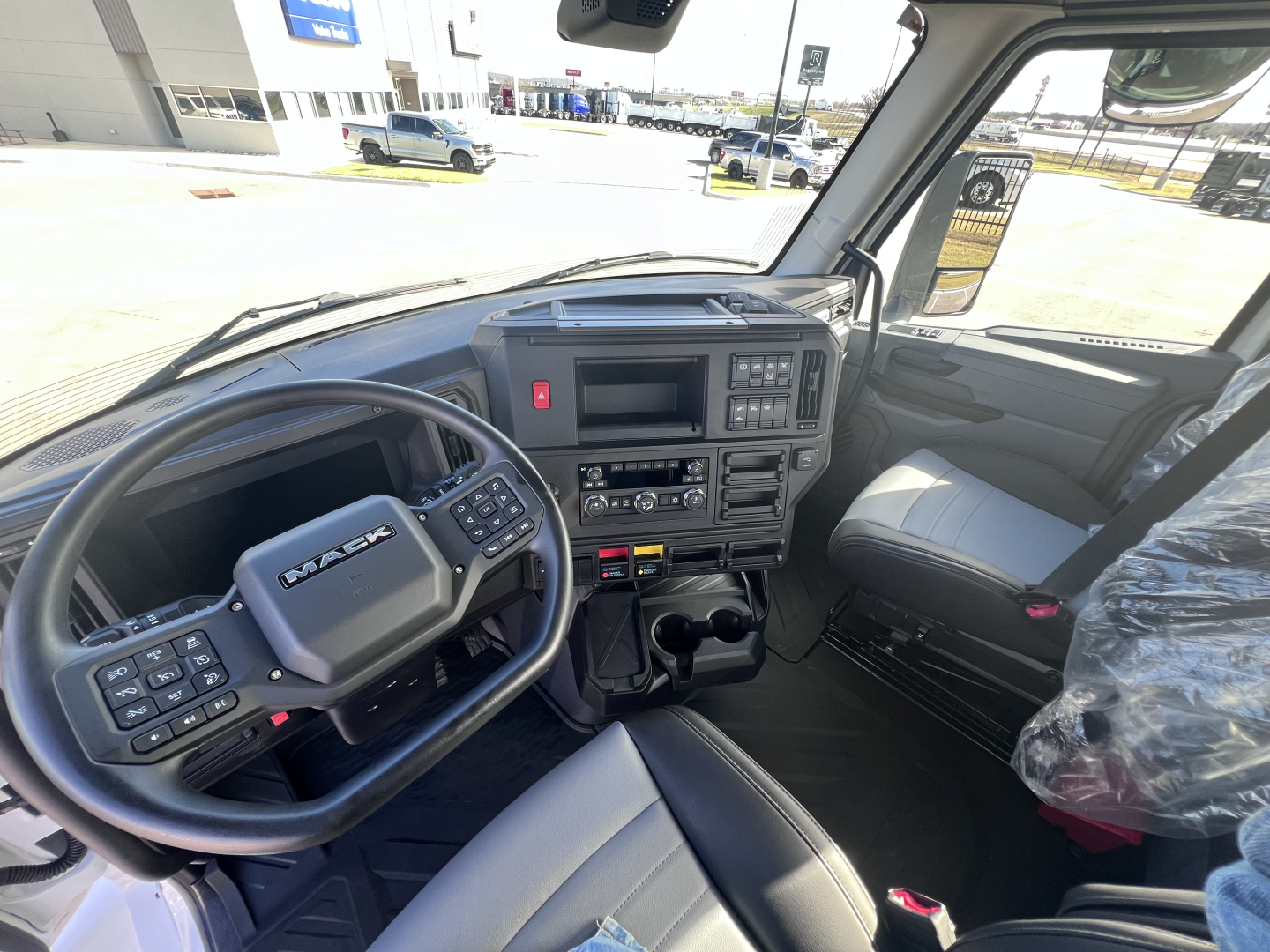 Truck cabin interior of a 2027 Mack AN64T showing steering wheel, dashboard with controls, gear shifter, and driver seat; side window and windshield visible.
