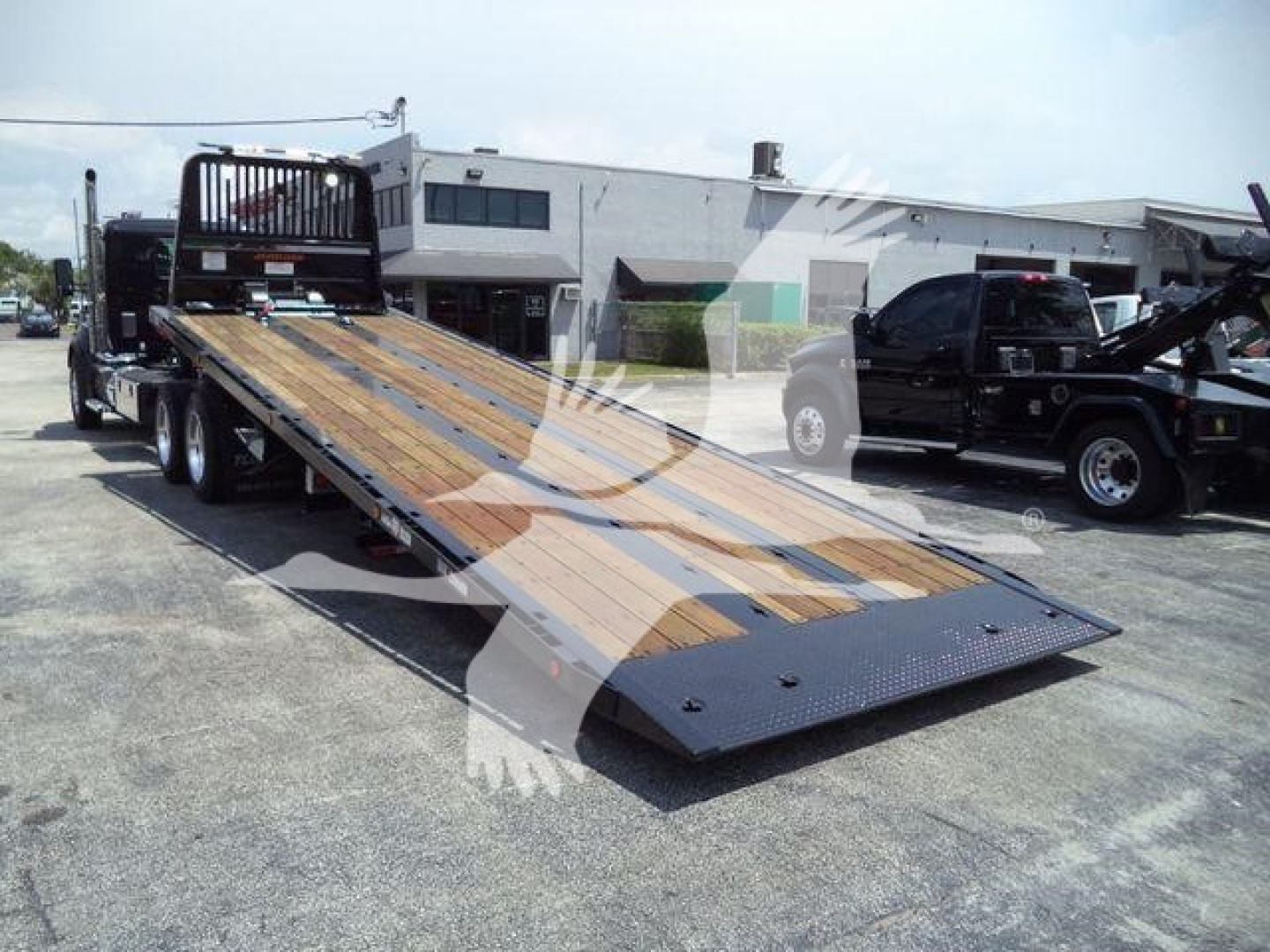 Kenworth truck bed with a rollback flatbed trailer on a loading lot, showing a wooden planked deck and black steel ramp end.