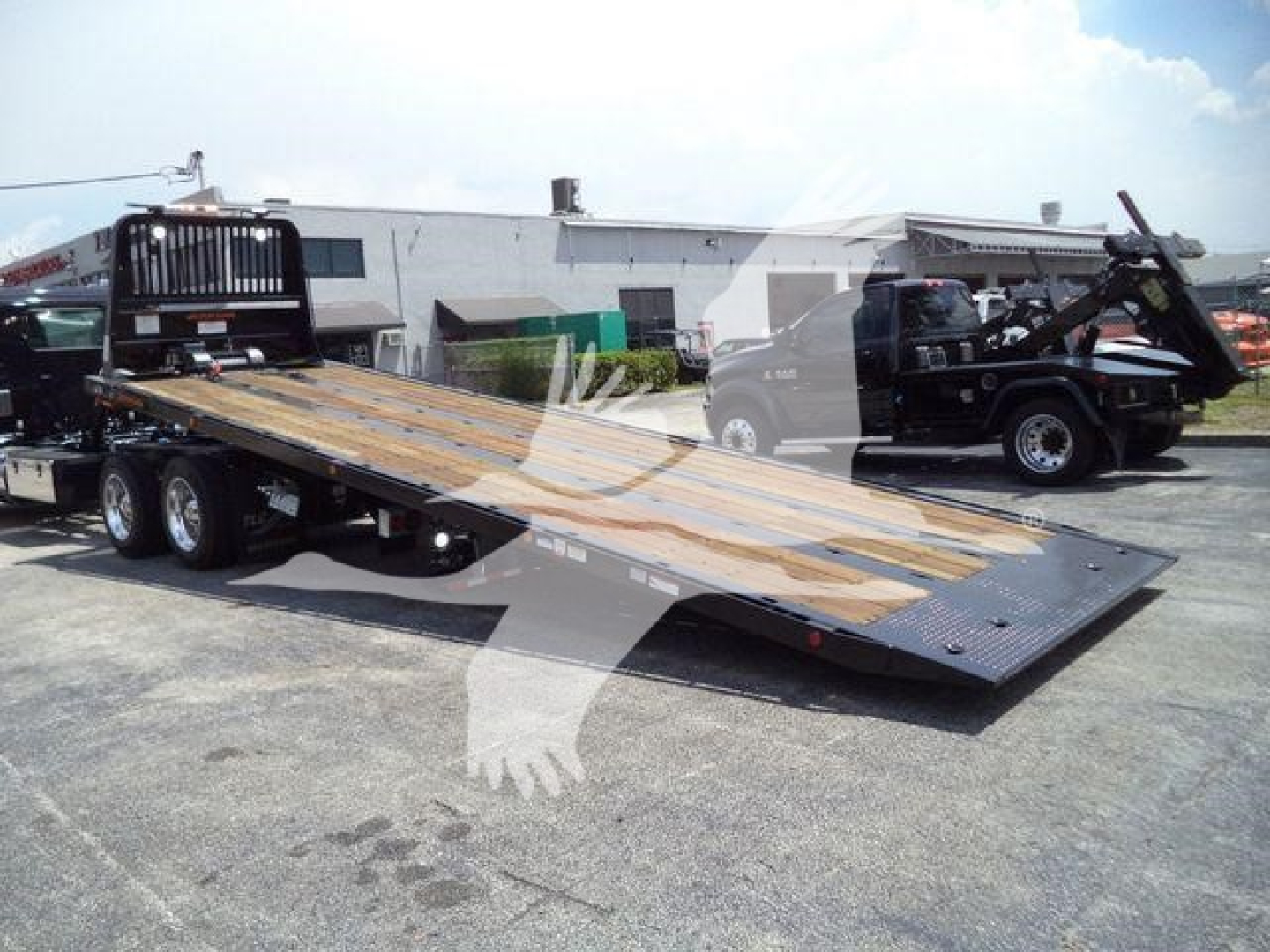 Kenworth T880 rollback truck with flatbed trailer and wooden planks, side view showing dual rear axles, metal ramps, and loading equipment in a lot.