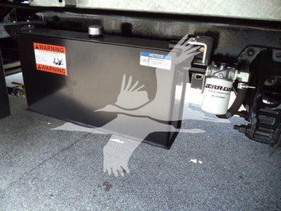 Underneath a Kenworth T880 showing a large black battery box with orange WARNING labels, a white logo of a bird overlay, and a white/blue notice sticker.