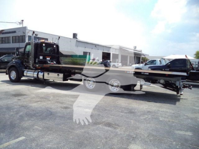 Kenworth T880 rollback tow truck with flatbed trailer and hydraulics, black cab, trailer with wheel chocks, dealership lot in background.