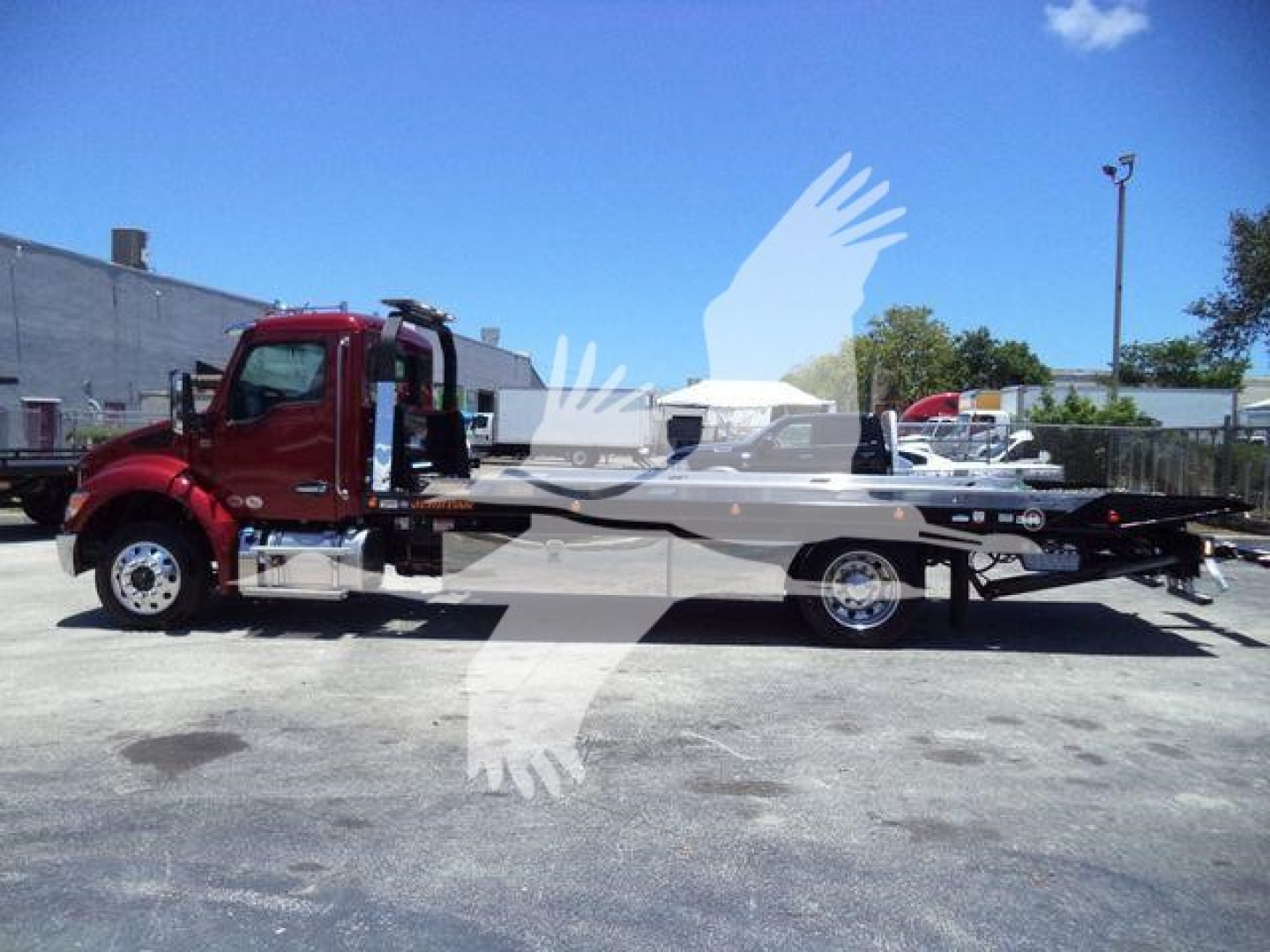 Kenworth T280 rollback tow truck in a parking lot, red cab with long flatbed, chrome mirrors and wheels visible.