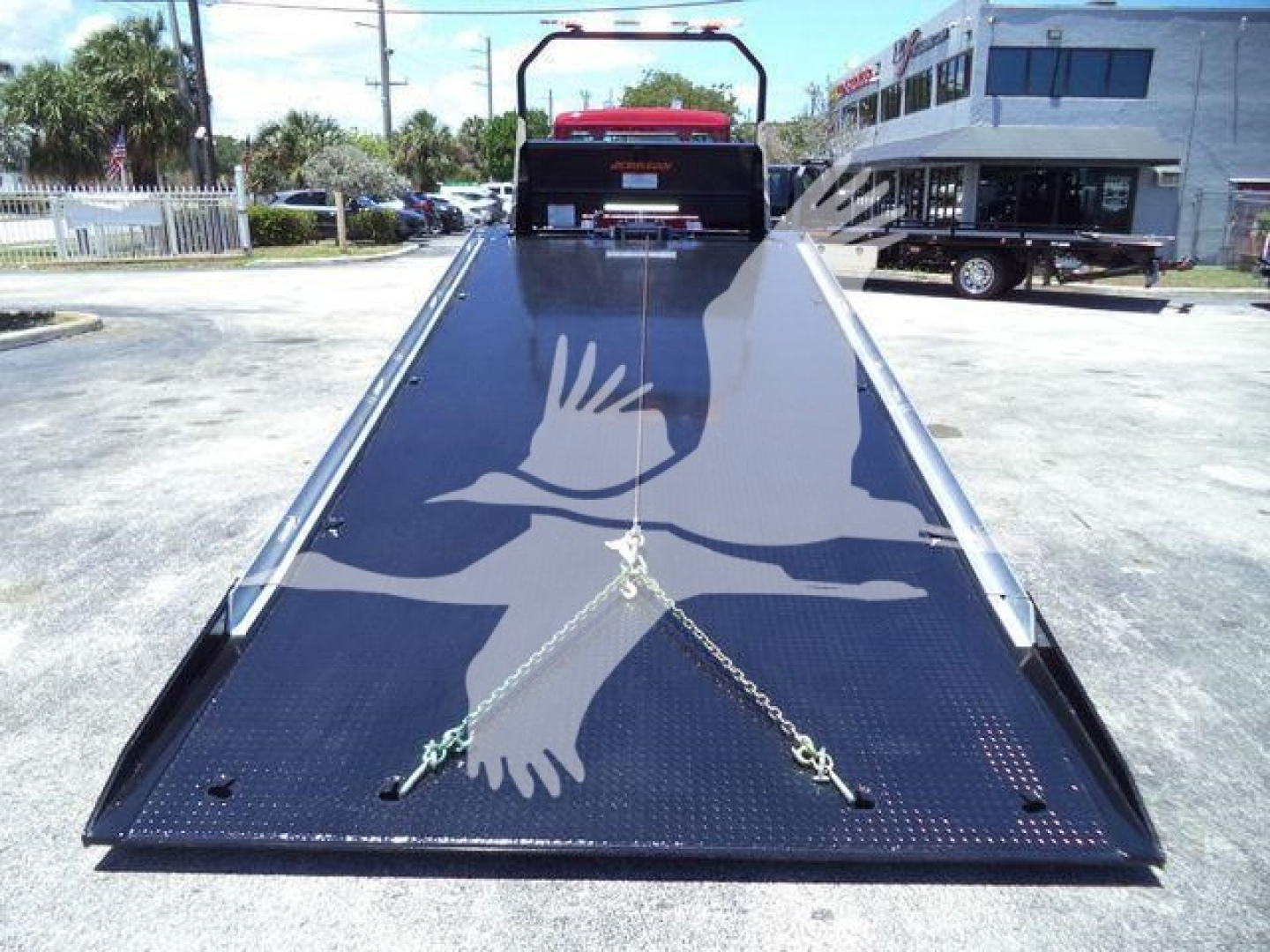 Truck bed with a hydraulic rollback flatbed extended, chains securing the ramp, Kenworth truck at back in outdoor lot.