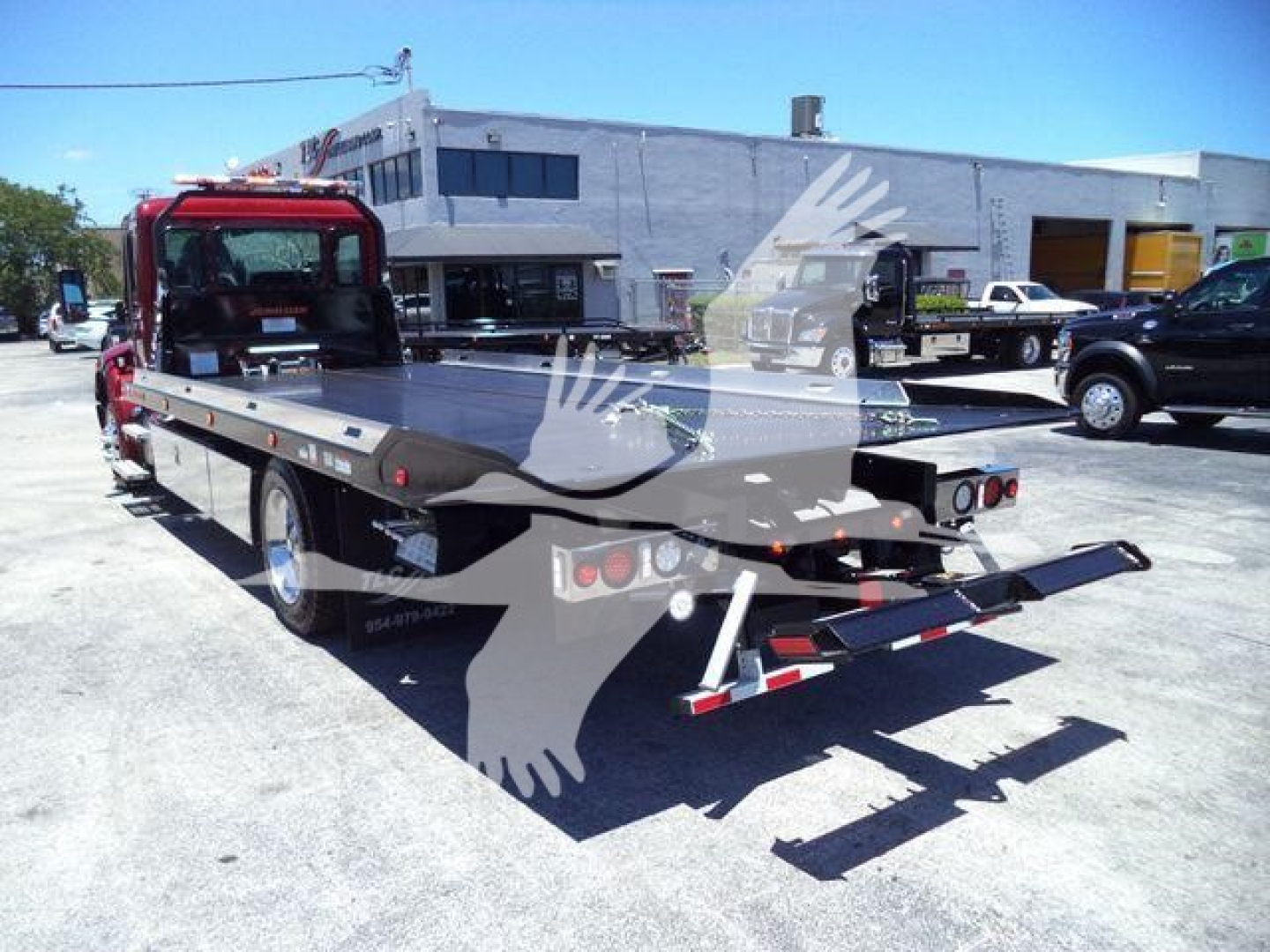 Flatbed tow truck with rear ramp and hitch, red cab, large flatbed, rollout equipment in lot; some trucks and a building in background.