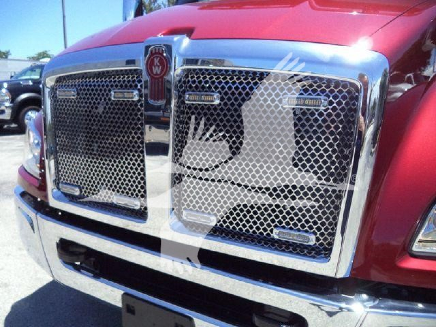 Front grille of a red Kenworth T-series truck with chrome mesh grille and KW emblem; partial view of bumper and hood; badge text not fully legible.