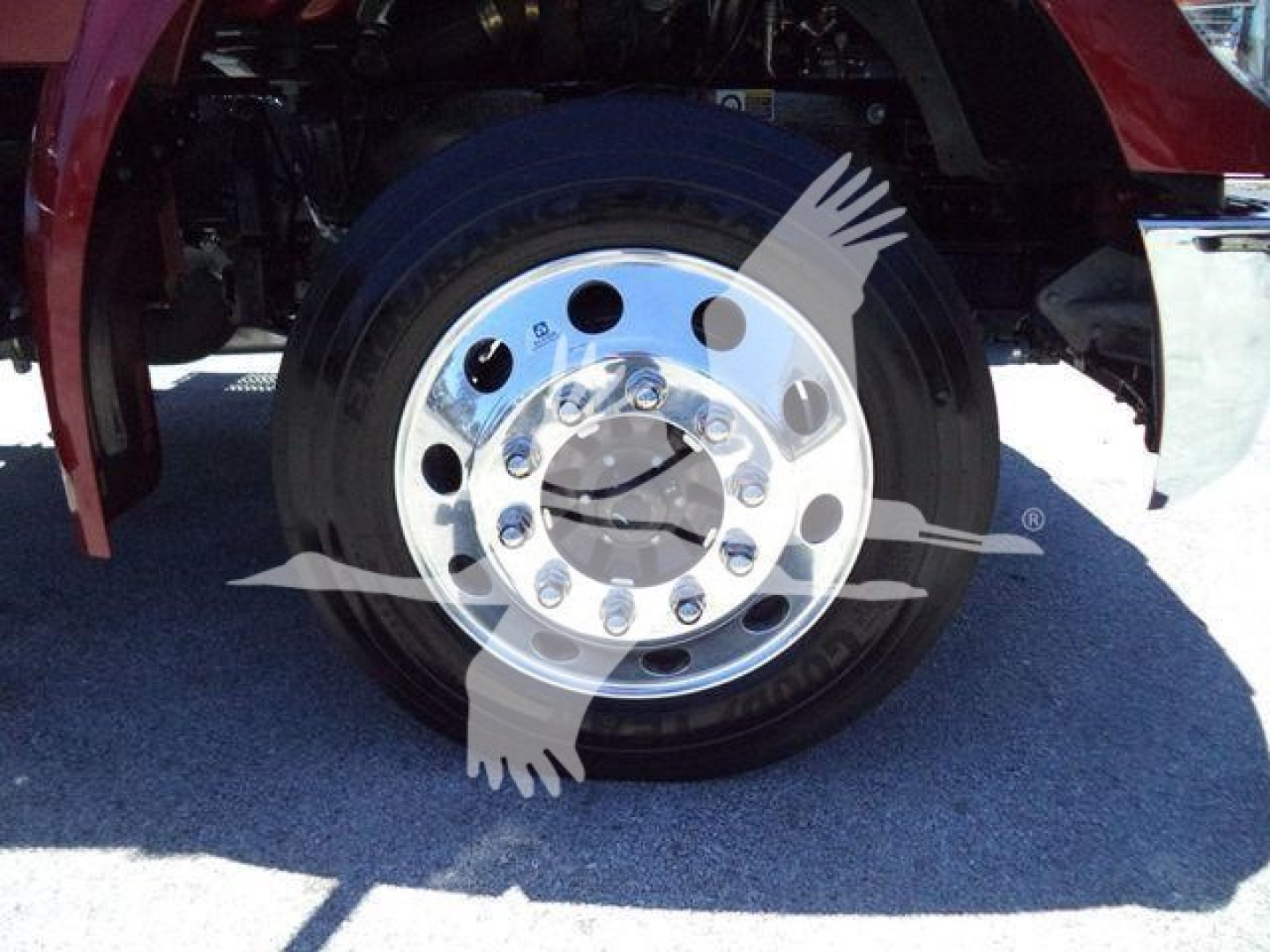 Truck wheel and chrome rim close-up showing front axle with black tire and polished steel hub, portion of red fender and shadowed ground.