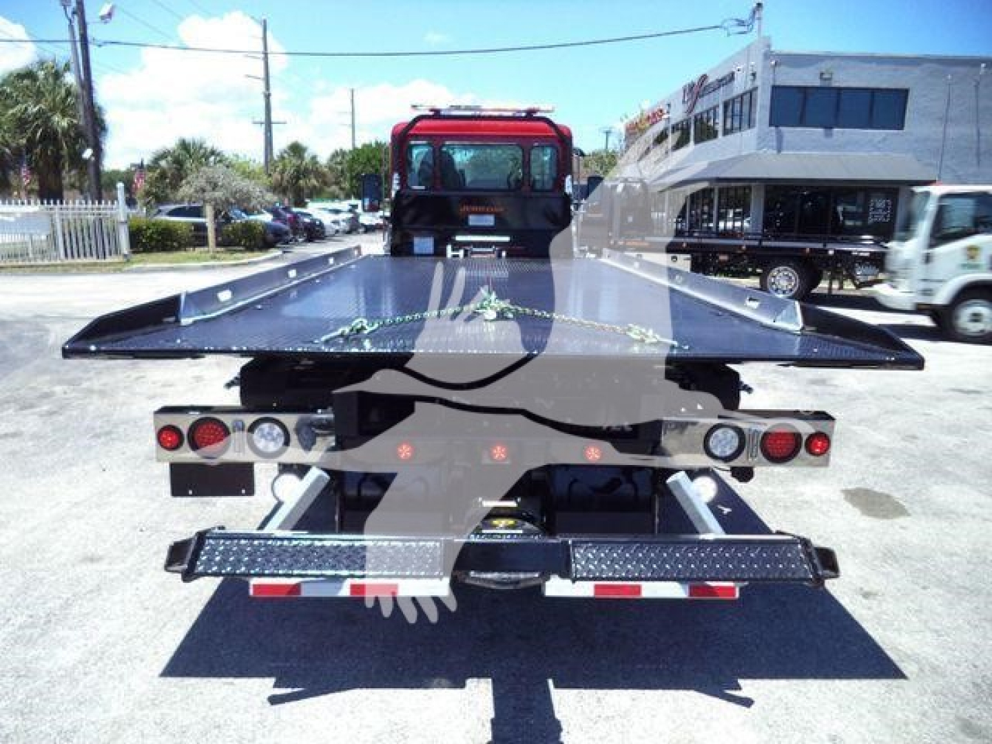Rear view of a tow truck flatbed rollback with a black metal bed extended, dual rear lights, and a red cab in the background.