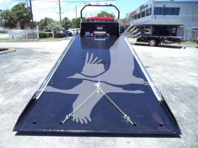 Truck bed with a hydraulic rollback flatbed extended, chains securing the ramp, Kenworth truck at back in outdoor lot.