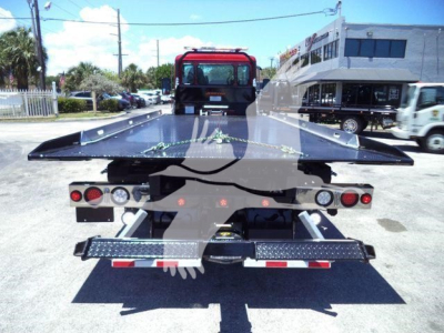Rear view of a tow truck flatbed rollback with a black metal bed extended, dual rear lights, and a red cab in the background.