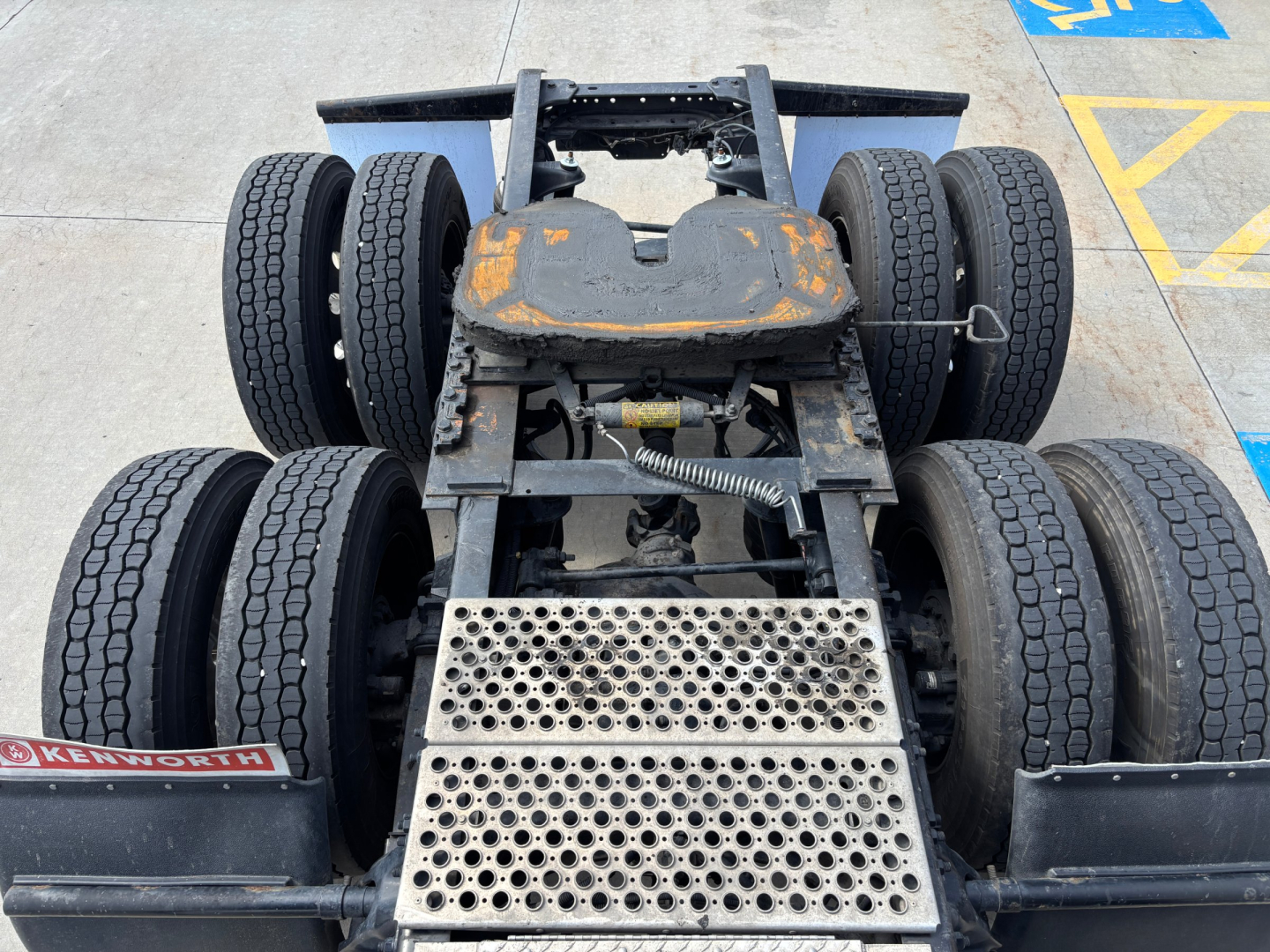 Kenworth T680 chassis showing rear view with dual rear axles and flatbed feeder plate, faded orange paint on top of fifth wheel, visible suspension and tires.