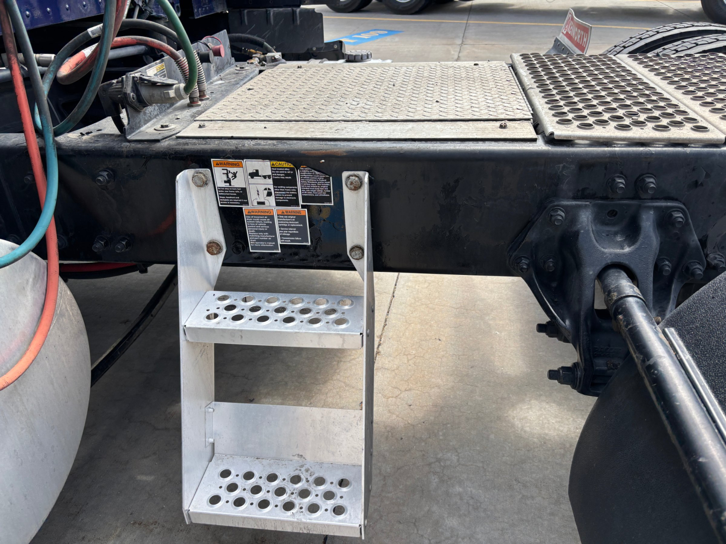 Kenworth T680 chassis with metal step ladder attached under cab, fuel lines and hydraulic components visible, VIN tag and warning labels on side of frame.