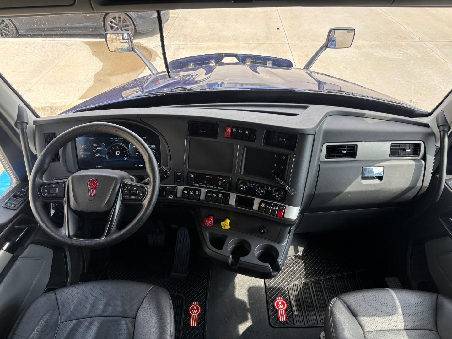 Driver cockpit of a Kenworth truck featuring a large steering wheel with red logo, multiple gauges, central control panel, and two cup holders. Mirrors visible through windshield.