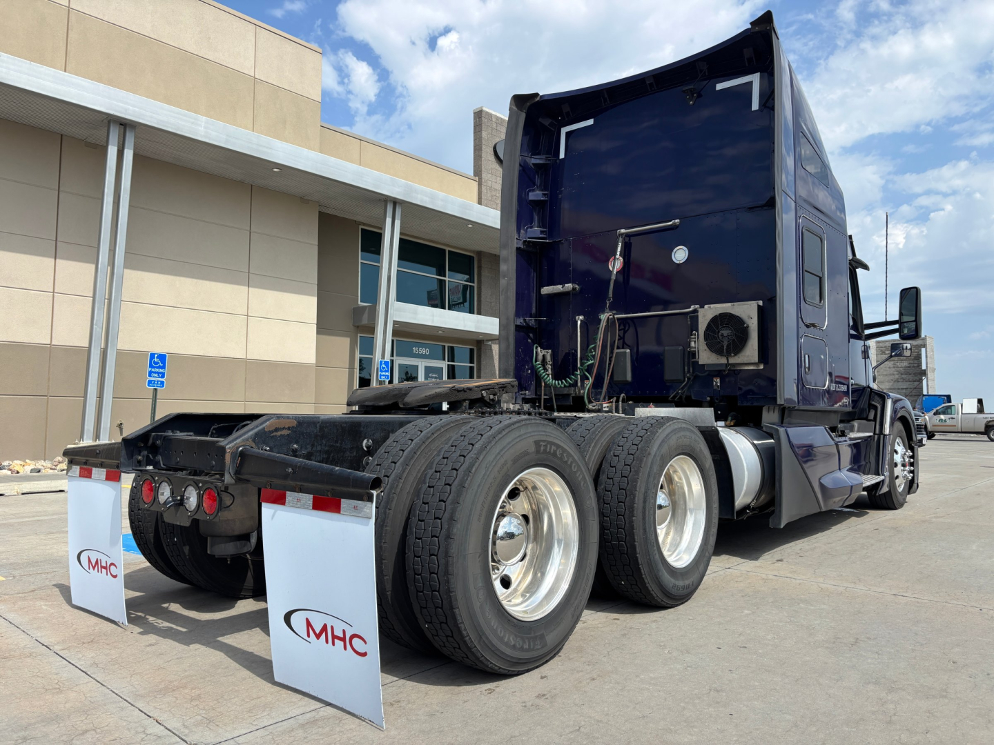Kenworth T680 tractor rear view showing dual rear axles and fifth wheel, with MHC mud flaps and blue cab; visible tires and chassis in a parking lot.