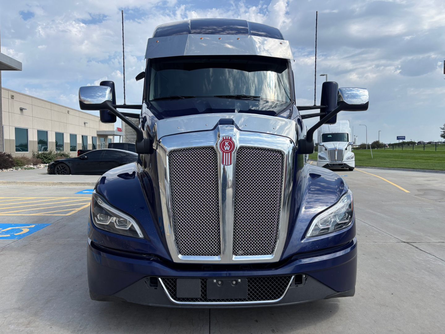 Front view of a 2024 Kenworth T680 truck showing chrome grille, large emblem, side mirrors, and headlights in a parking lot.