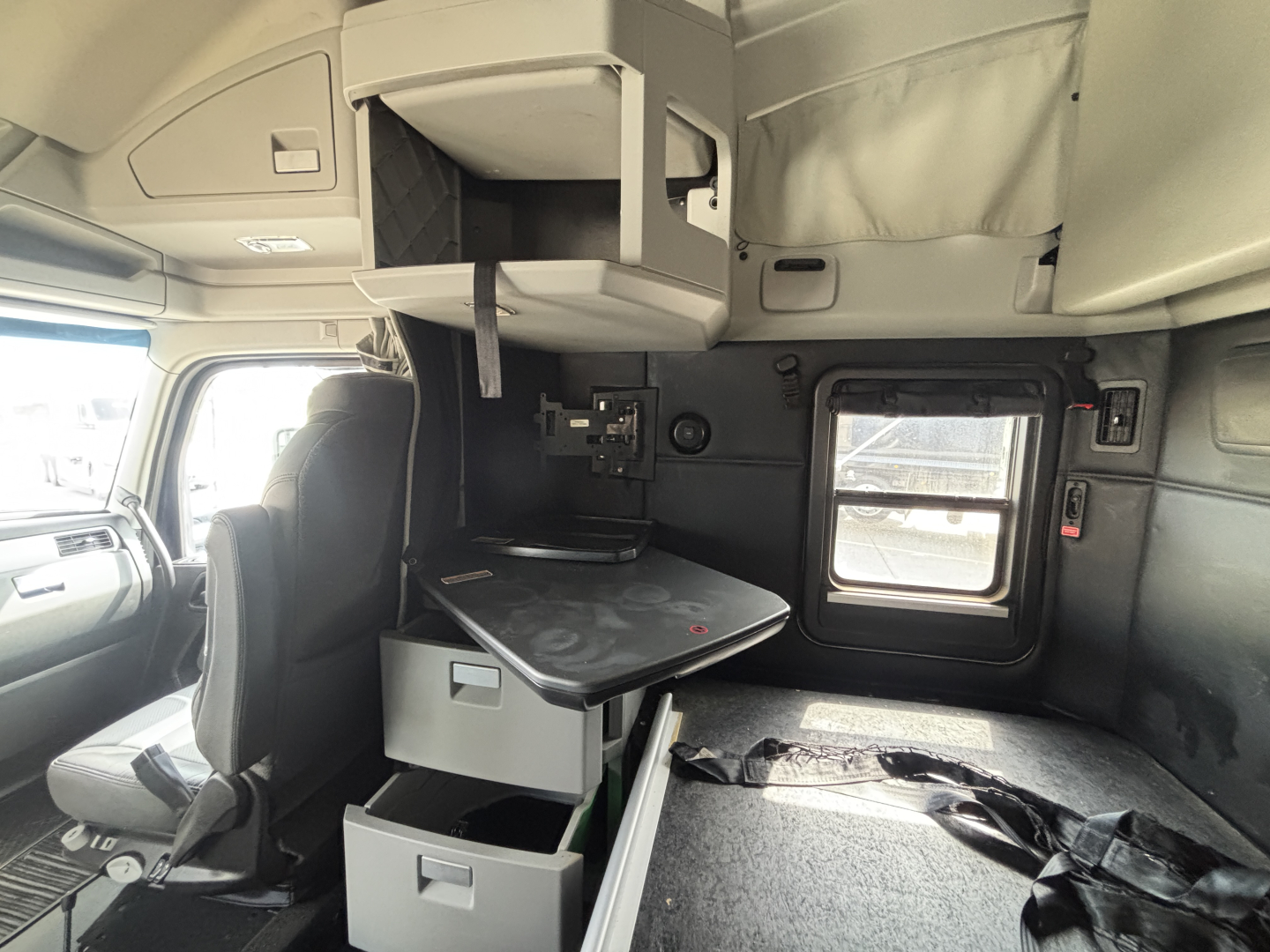 Cab interior of a 2024 Kenworth T680 showing sleeper cab with overhead storage, small desk, drawers, and window; dashboard and driver's seat visible.