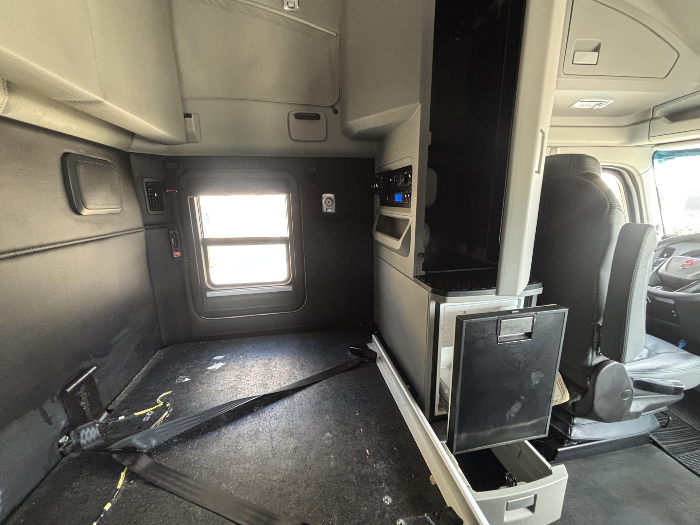 Kenworth T680 truck cabin interior showing sleeper area with small window, storage cabinets, and driver seat; floor area with seat belt and dashboard visible.