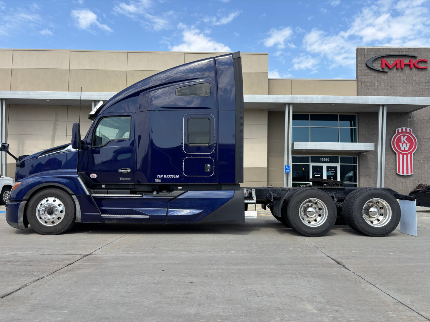 Kenworth T680 side view showing blue semi truck with sleeper cab and dual rear axles in showroom lot.