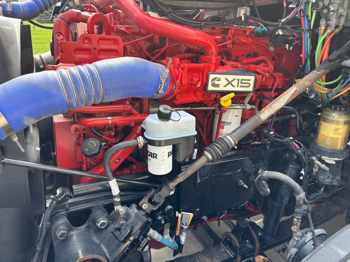 Kenworth T680 engine bay showing X15 badge, red engine, blue intercooler piping, oil reservoir, and various hoses and wiring.
