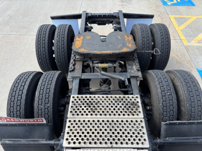 Kenworth T680 chassis showing rear view with dual rear axles and flatbed feeder plate, faded orange paint on top of fifth wheel, visible suspension and tires.