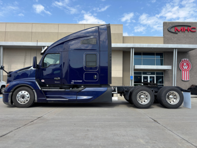 Kenworth T680 side view showing blue semi truck with sleeper cab and dual rear axles in showroom lot.