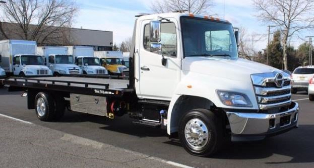 Flatbed tow truck showing front grille, white cab, dual mirrors, chrome bumper and flatbed with wheel chocks.