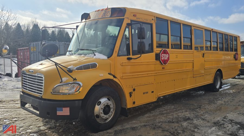 School bus in-service with yellow body and stop sign extended on the side, front grille and headlights visible, Freightliner bus chassis, snow on ground.