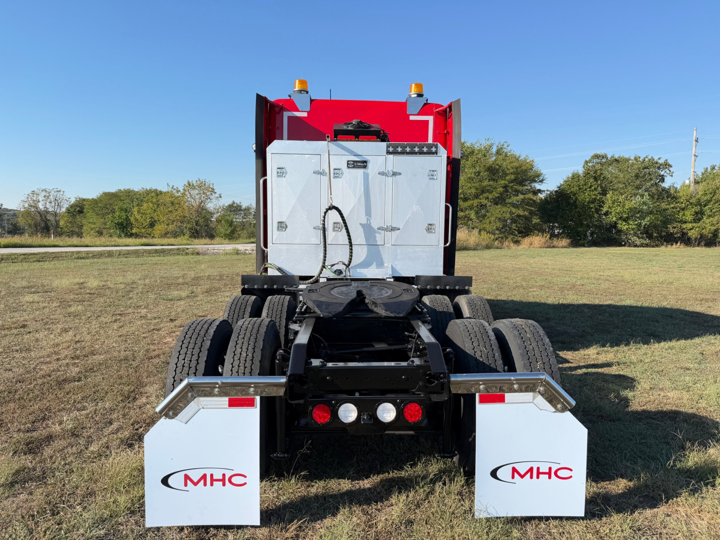 Kenworth truck rear view showing dual rear wheels and metal toolbox with 'MHC' logo on white mud flaps.