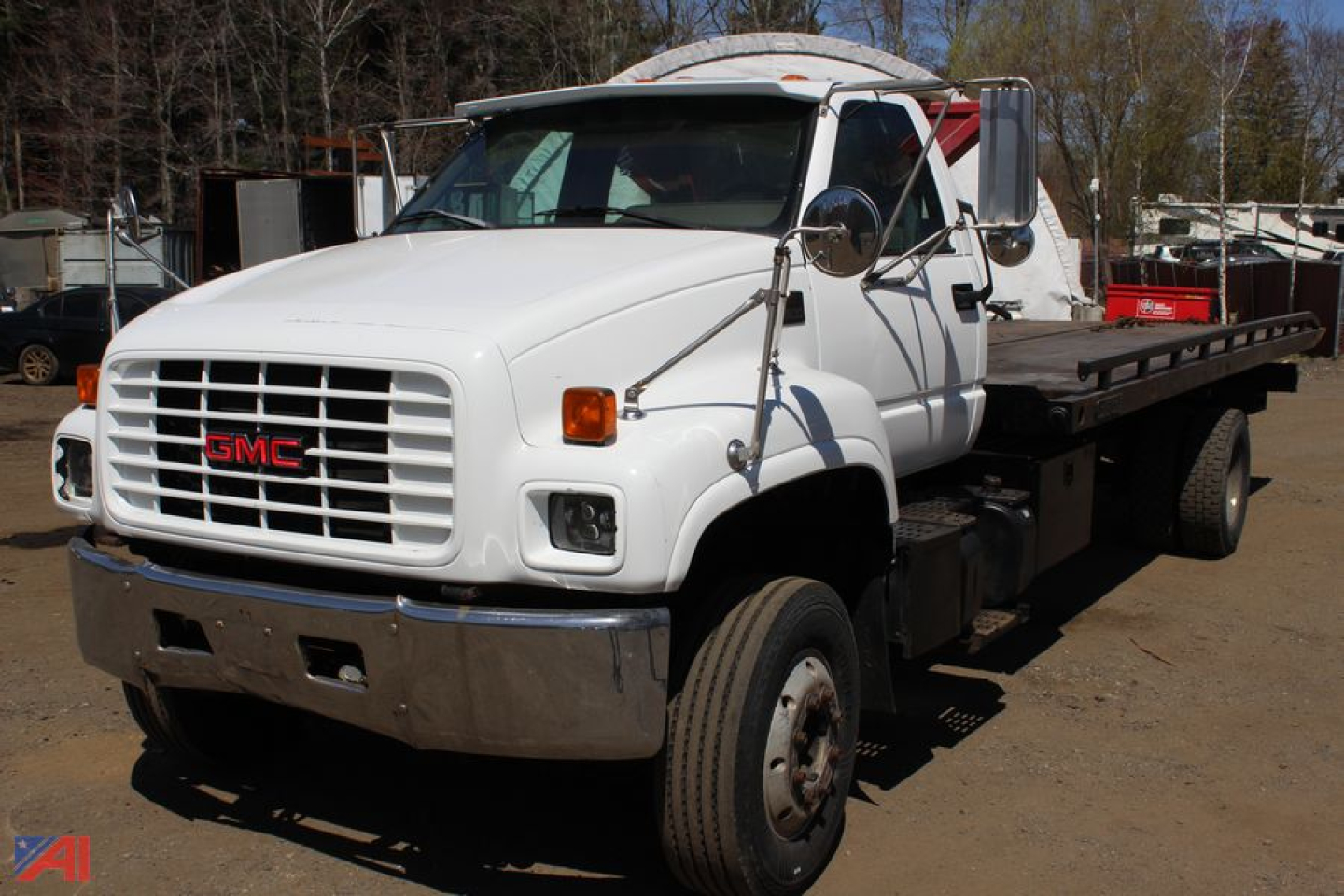 GMC medium-duty tow truck / carrier with flatbed rear, white cab, big chrome bumper and GMC grille, side mirrors, and dual rear wheels in outdoor lot.