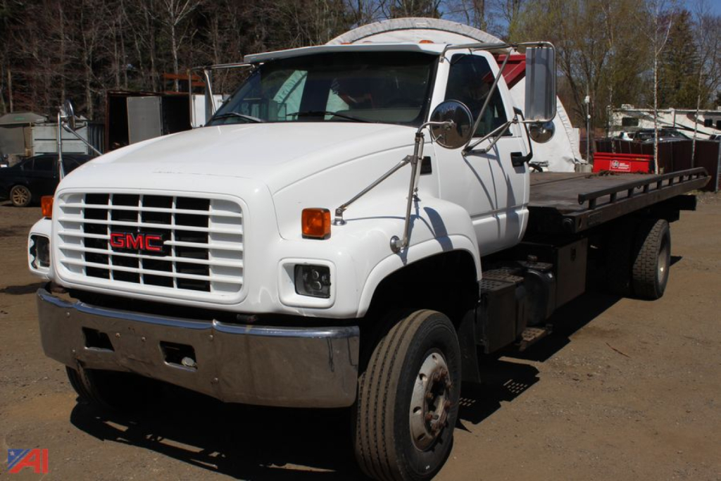 GMC medium-duty tow truck / carrier with flatbed rear, white cab, big chrome bumper and GMC grille, side mirrors, and dual rear wheels in outdoor lot.