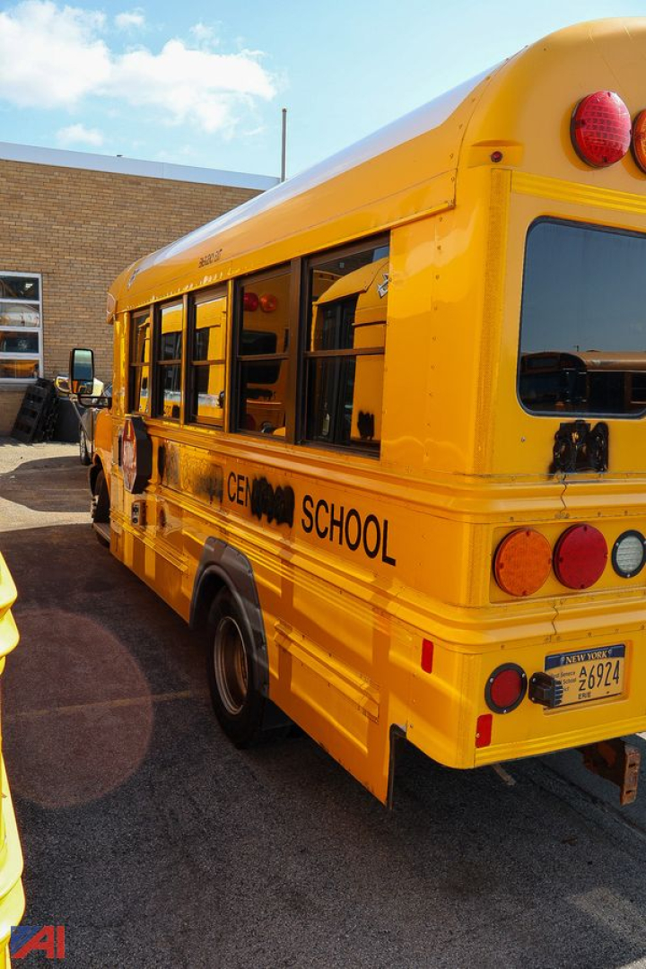 School bus rear-left side showing bright yellow body with 'SCHOOL' lettering and tinted windows; GEDN bus number plate visible; part of a brick building in background.