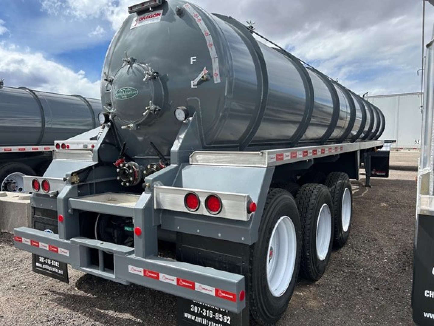 Trailer tank with cylindrical steel tank and triple axle wheels in a yard; rear shows taillights, bumper, and safety decals; circular hatch and inspection ports on tank.