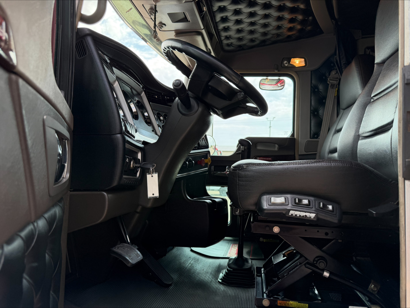 Kenworth W900L cab interior showing steering wheel, driver seat with controls, and dashboard in view.