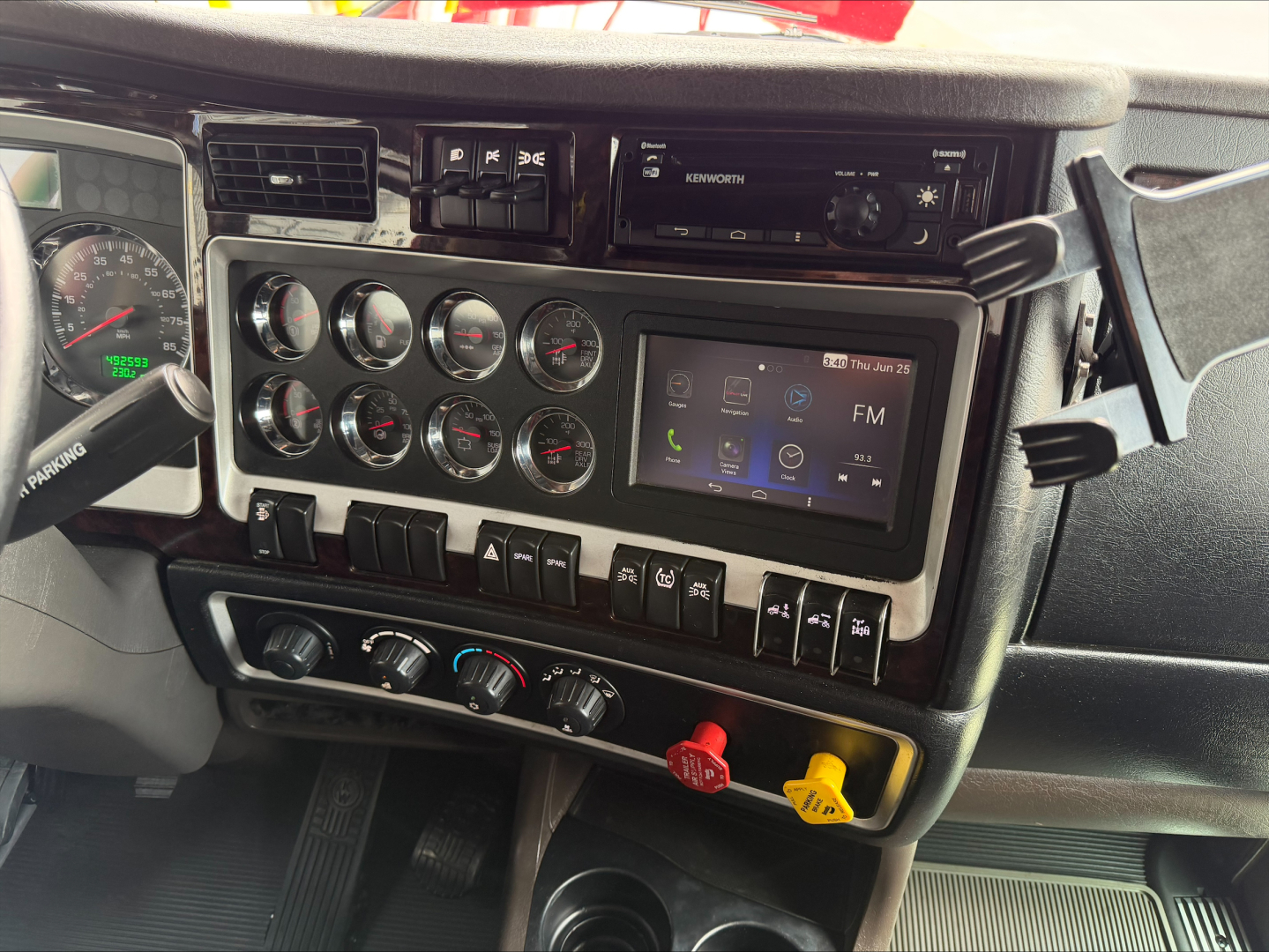 Kenworth W900L cab interior showing dashboard gauges, infotainment touchscreen, climate controls, and red/yellow knobs.