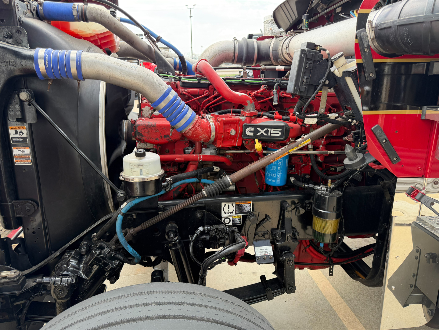 Kenworth W900L engine bay with red 15-liter engine, blue and red hoses, and various fluid reservoirs visible; intake and exhaust piping, belts, and wiring in view.