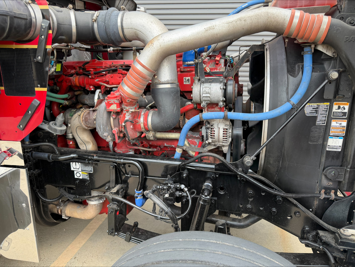 Kenworth W900L engine bay showing red engine block, silver intake pipes, blue hoses, and black chassis with various components.