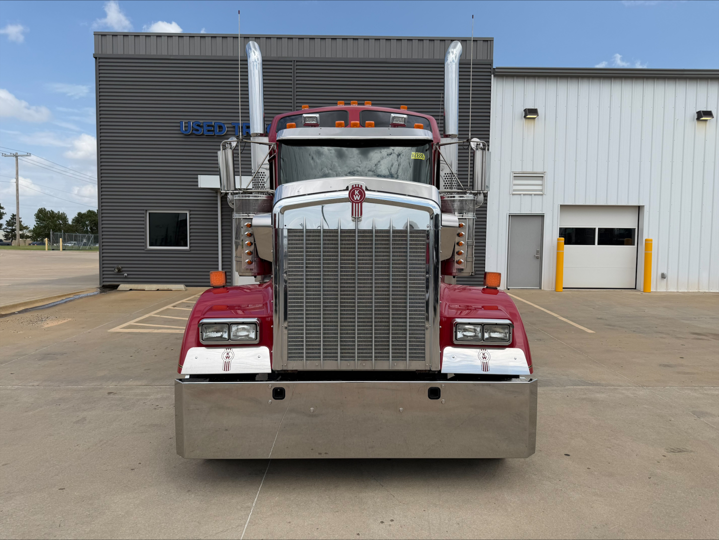 Kenworth W900L front view showing large chrome grille, dual exhaust stacks, and tall bumper with silver air intake; truck in front of a building with 'USED' sign visible.