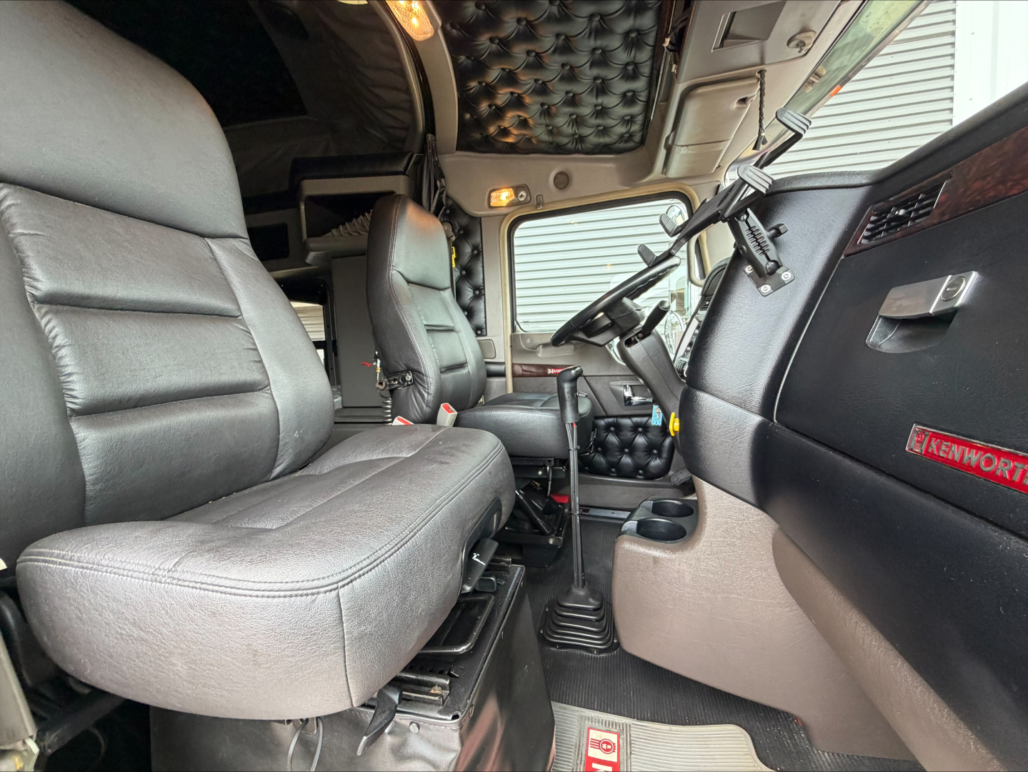 Kenworth W900L truck interior cab view showing driver seat and passenger seat with black leather and beige carpet, steering wheel, dashboard, and quilted headliner.