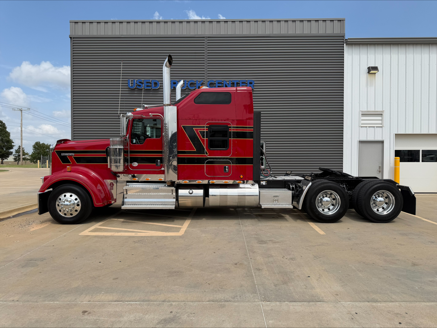 Kenworth W900L tractor in red and black, side view, with chrome fuel tanks and dual rear axles. Visible text includes 'USED TRUCK CENTER' on the building behind.
