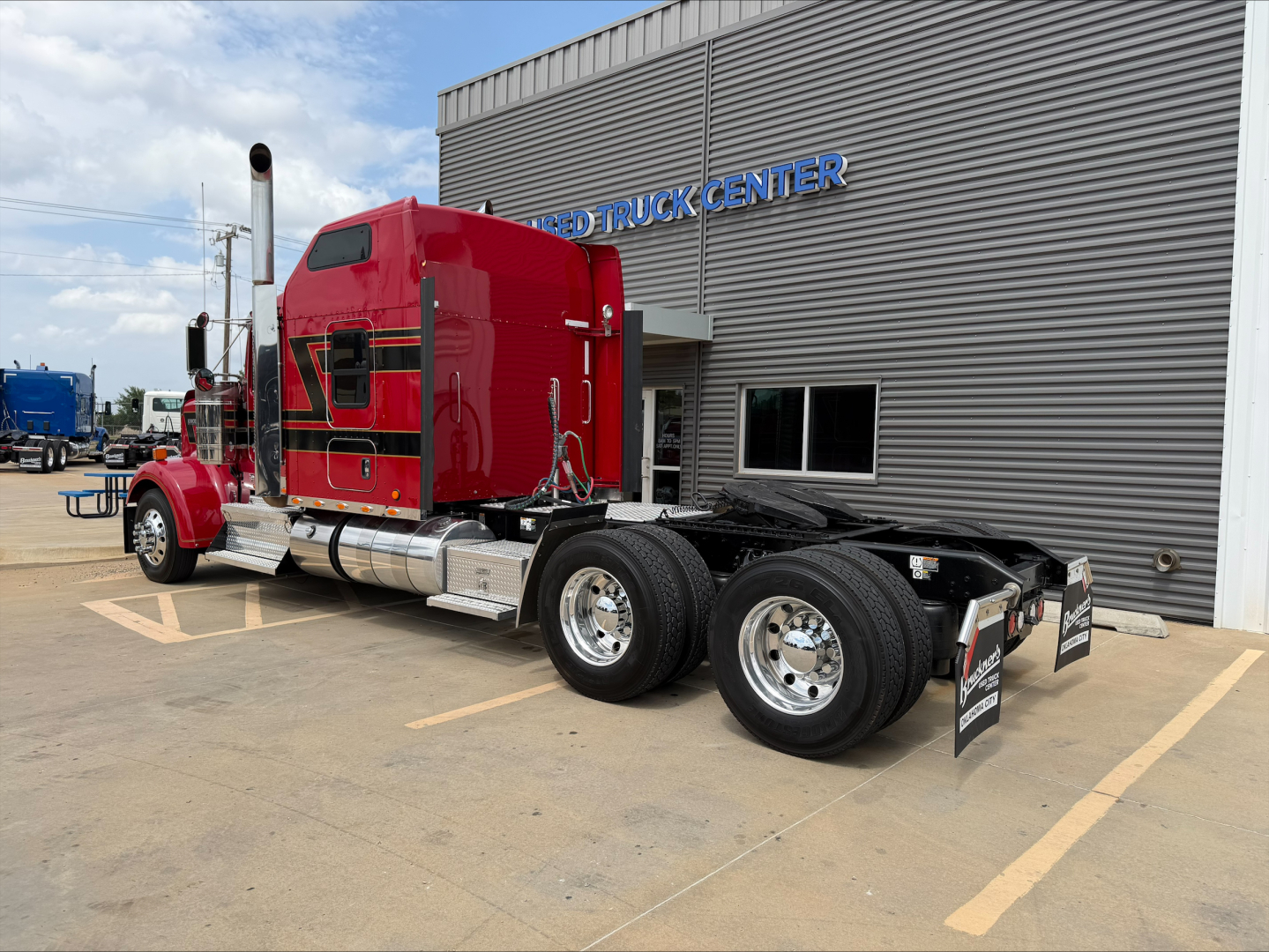 Kenworth W900L red diesel truck tractor in a dealership lot with chrome fuel tanks and dual rear axles, side view, adjacent building.