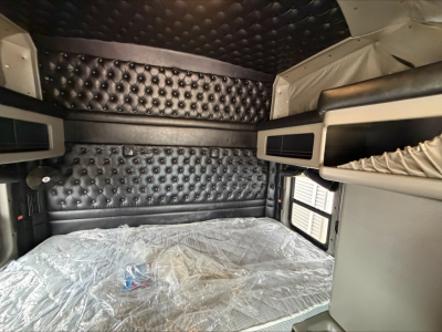 Kenworth W900L truck cab interior with quilted black cushions and bunk area. Visible plastic-wrapped mattress. Side window and storage compartments.