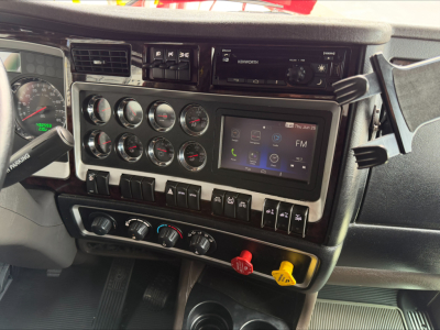 Kenworth W900L cab interior showing dashboard gauges, infotainment touchscreen, climate controls, and red/yellow knobs.