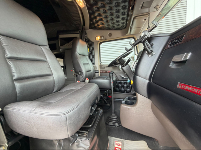 Kenworth W900L truck interior cab view showing driver seat and passenger seat with black leather and beige carpet, steering wheel, dashboard, and quilted headliner.