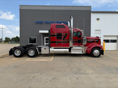 Kenworth W900L tractor in red with black chassis, side view showing cab logo and chrome fuel tanks; dealership building in background reads 'USED TRUCK CENTER'