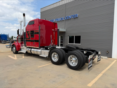 Kenworth W900L red diesel truck tractor in a dealership lot with chrome fuel tanks and dual rear axles, side view, adjacent building.