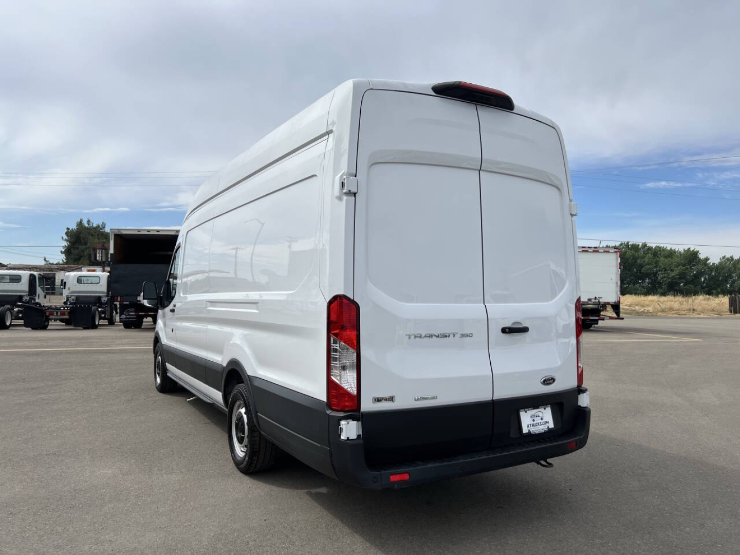 Ford Transit van rear-side view showing cargo van with white exterior, black bumper, high roof, Ford badge, and rear doors; no visible text legible from this angle.